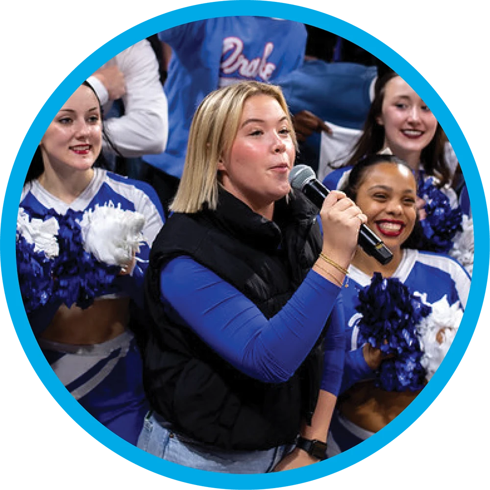 Mel Dygert speaks into a microphone at an event, wearing a blue long-sleeve shirt and black vest, with cheerleaders holding pom-poms in the background.