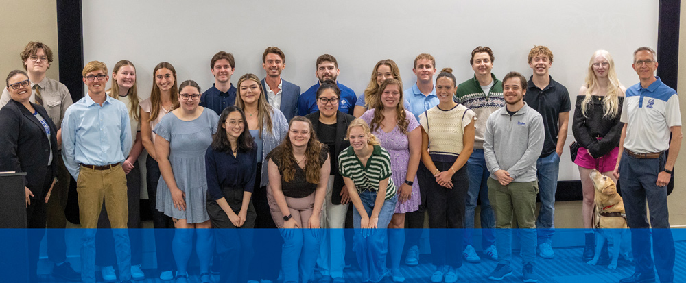 A group photo of people consisting of students and an instructor standing in front of a large projection screen in a lecture hall; A yellow lab service dog is sitting near the instructor on the far right