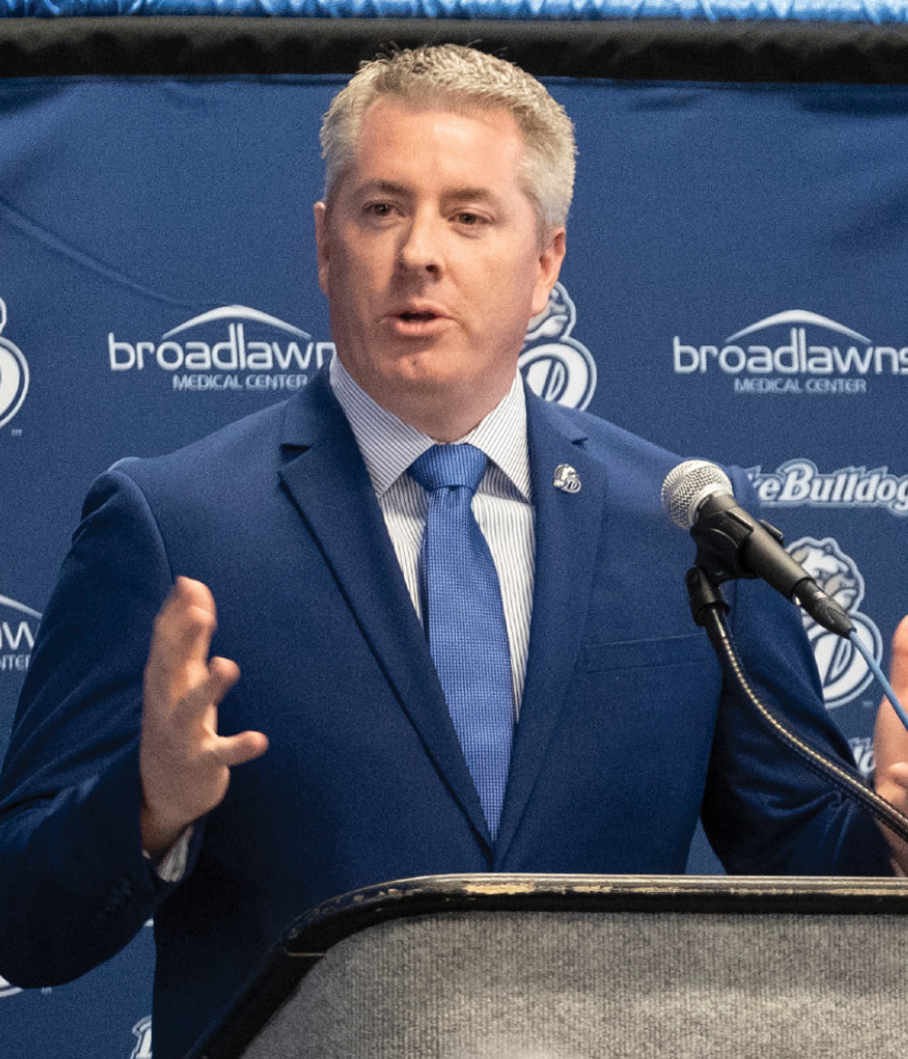A man with graying hair in a blue suit and tie gestures while speaking at a podium. The background is a blue banner featuring the Broadlawns Medical Center and Drake Bulldogs logos.