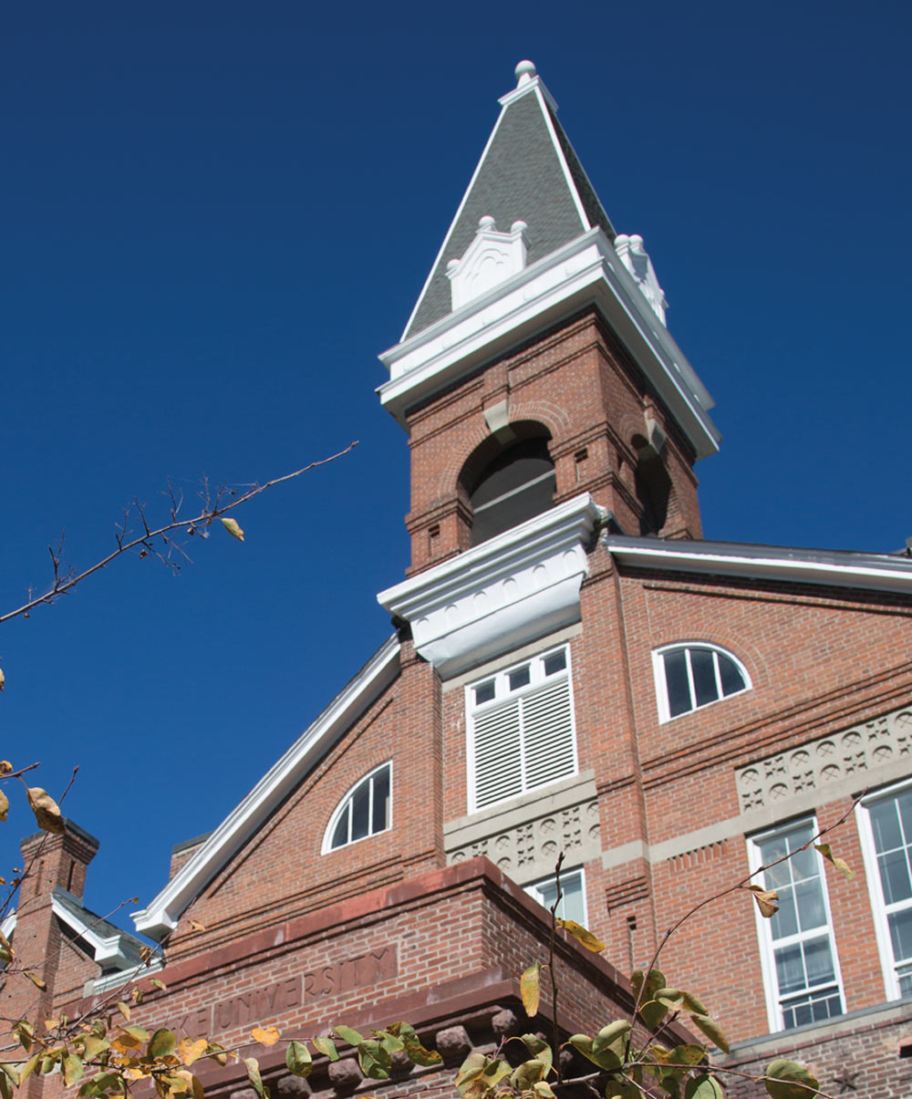 A low-angle view of a historic red brick building at Drake University with a prominent white and gray clock tower steeple set against a clear, vibrant blue sky.