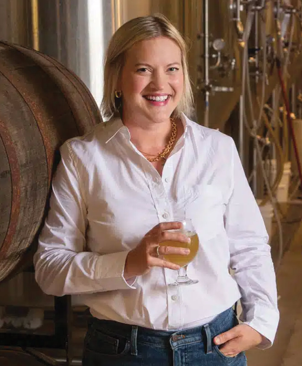 A smiling woman with blonde hair stands in a brewery wearing a white button-down shirt, holding a glass of light-colored beer. Large wooden barrels are visible behind her.