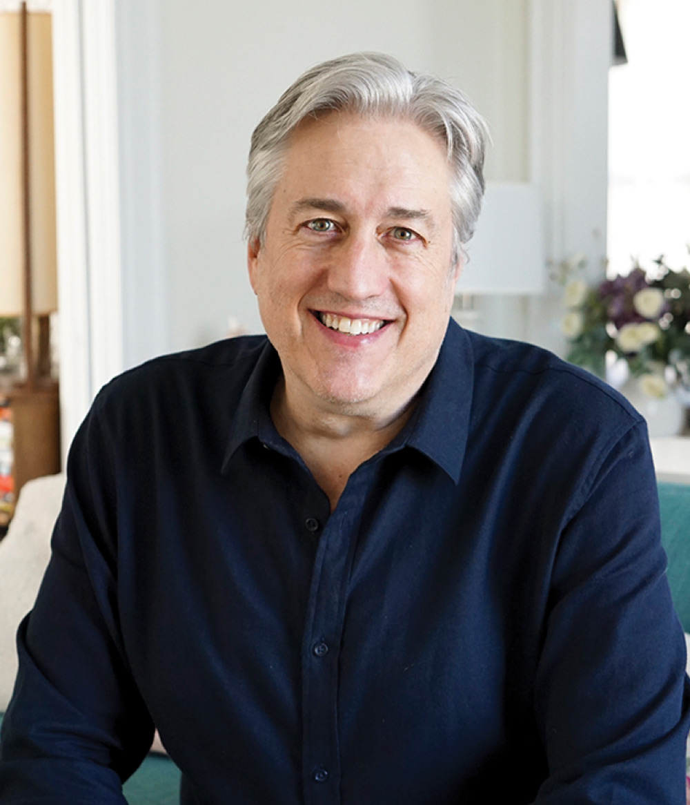 A close-up portrait of a smiling man with gray hair wearing a dark blue button-down shirt. The background is a bright, softly blurred indoor living space with flowers.