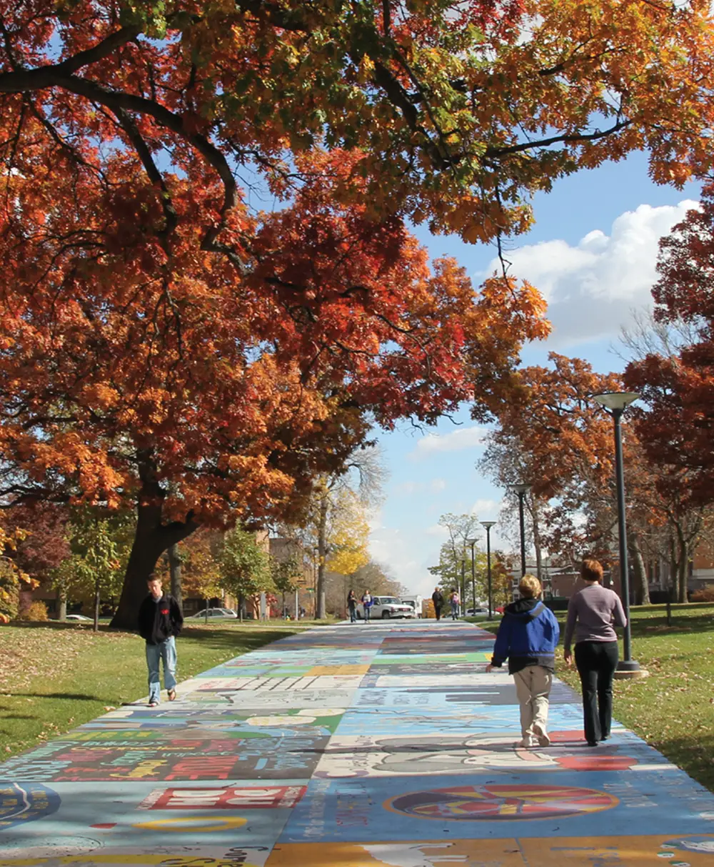 A wide campus walkway painted with colorful squares and murals. People walk toward the horizon under large trees with vibrant orange and red autumn leaves under a blue sky.