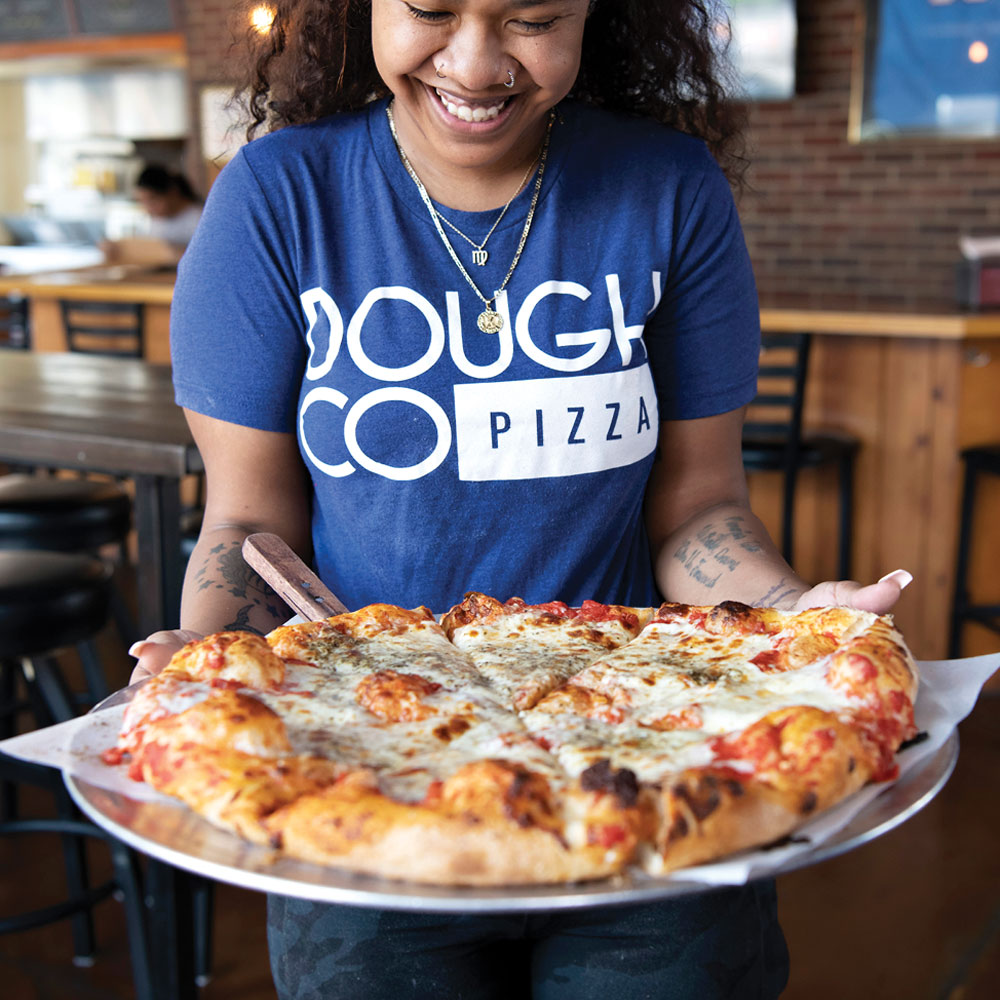 A staff member wearing a "Dough Co." shirt holding a fresh, hand-tossed pizza, showcasing one of the area's favorite casual eats.
