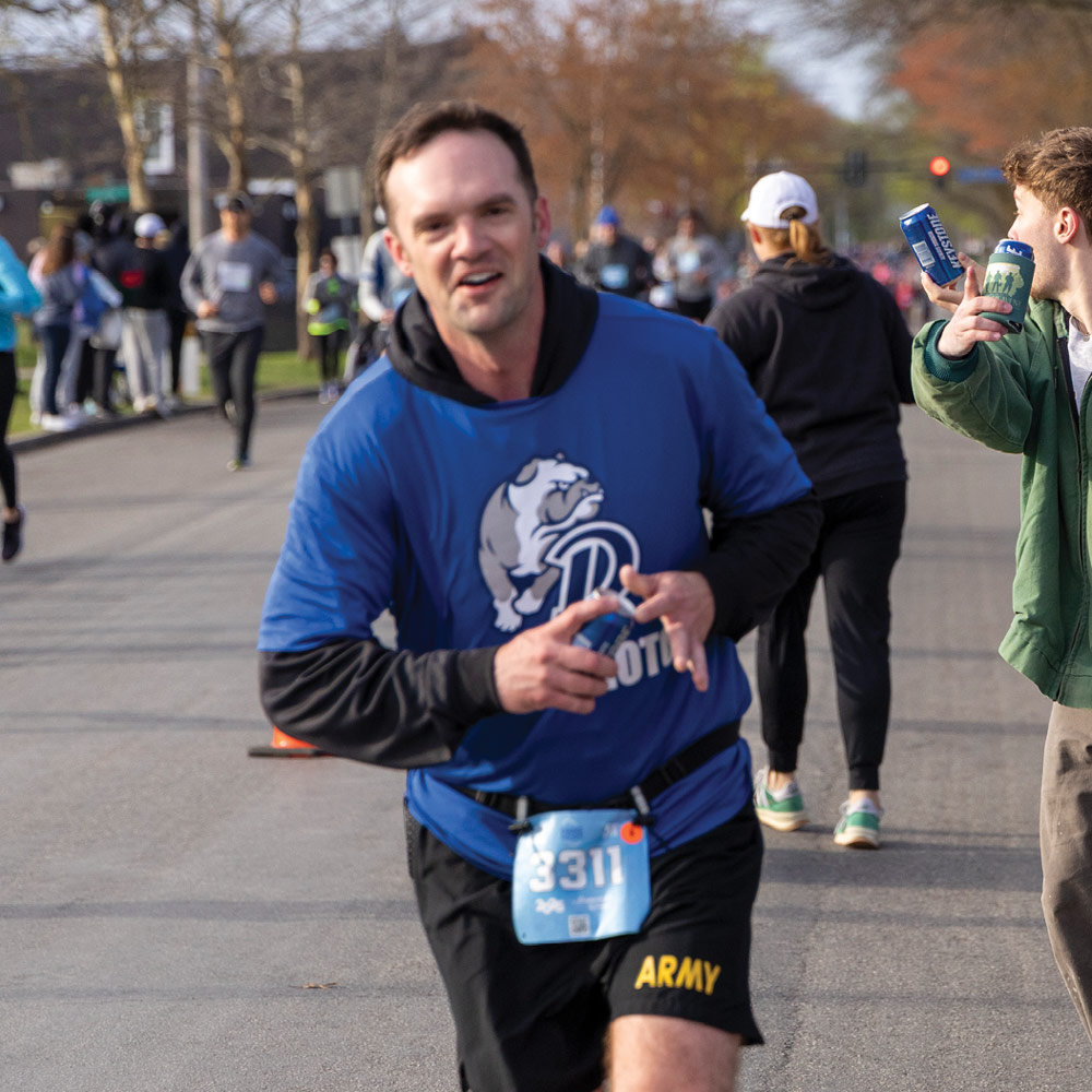 A man in a blue Drake University t-shirt with a bulldog mascot logo runs down a street during a road race. He holds a blue beverage can as a spectator cheers in the background.
