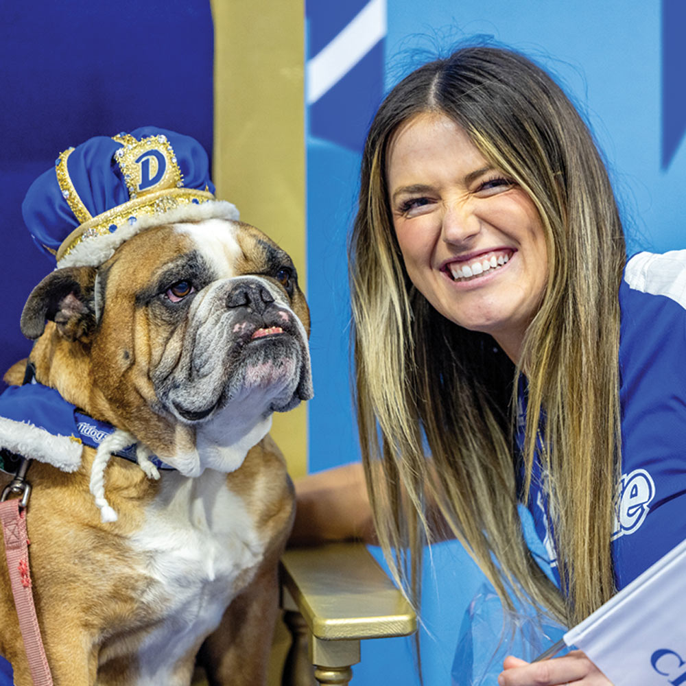 A smiling woman with long hair poses next to a bulldog mascot wearing a blue and gold crown.