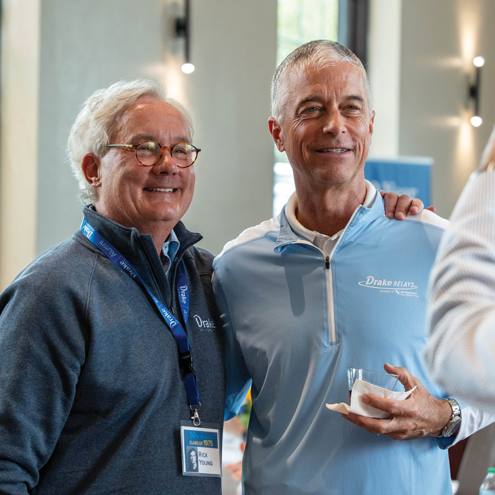 Two men, one in a grey quarter-zip and one in a light blue "Drake Relays" pullover, smile for a photo at an indoor event. The man on the left wears a "Rick Young" name tag.