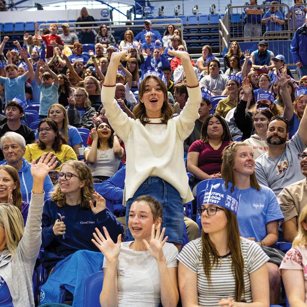 A crowd of spectators in stadium bleachers raises their hands in the air. A young woman in a white sweater stands in the center, cheered on during the Beautiful Bulldog Contest.