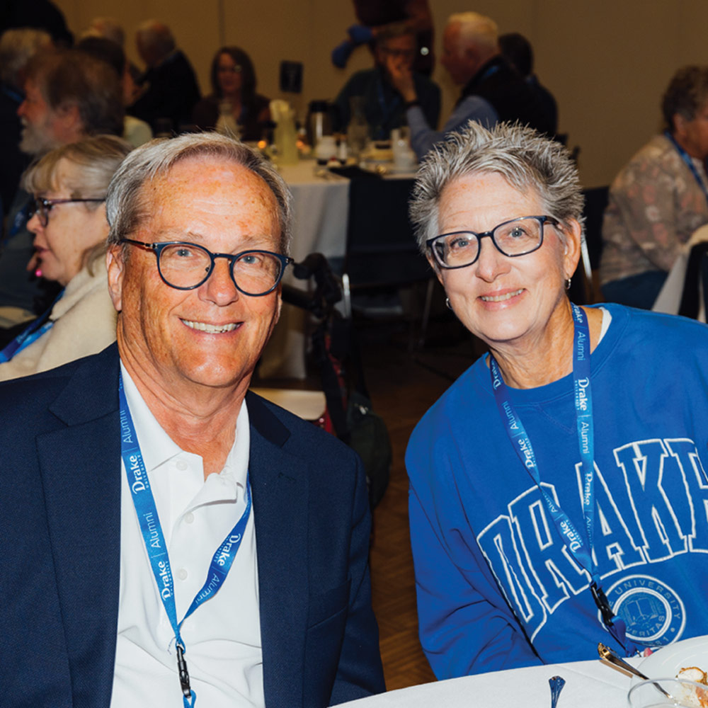 A man in a navy blazer and a woman in a blue Drake University sweatshirt smile for a close-up photo while seated at a banquet table. Both wear blue "Drake Alumni" lanyards.
