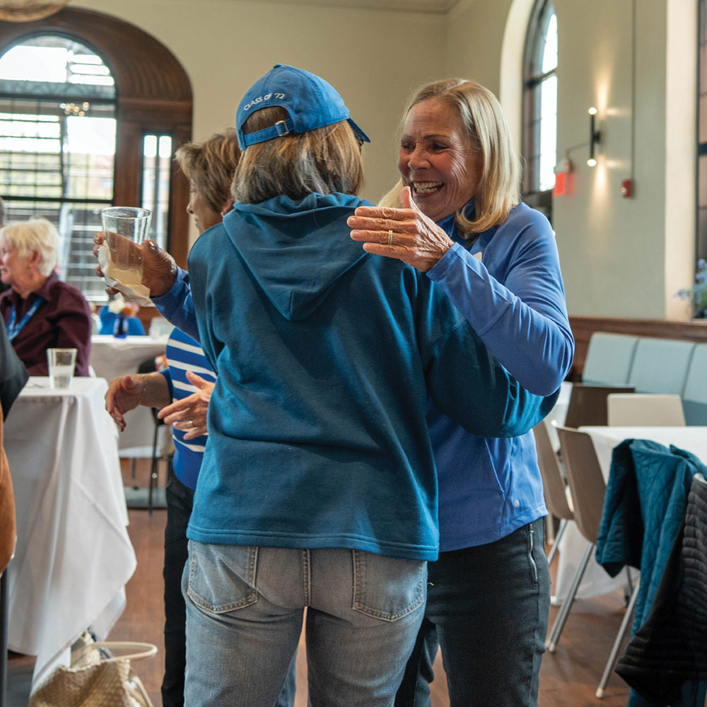 A woman in a light blue quarter-zip jacket smiles warmly while leaning in to hug another person wearing a dark blue hoodie and a "Class of 72" cap at an indoor reunion event.