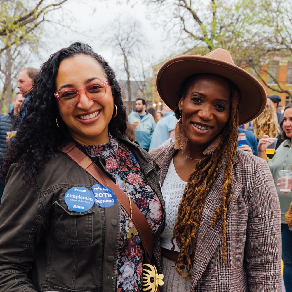 Two women smile for a photo at an outdoor reunion. One wears a brown hat and plaid blazer; the other has pink glasses and an "It's My 20th Reunion" button.