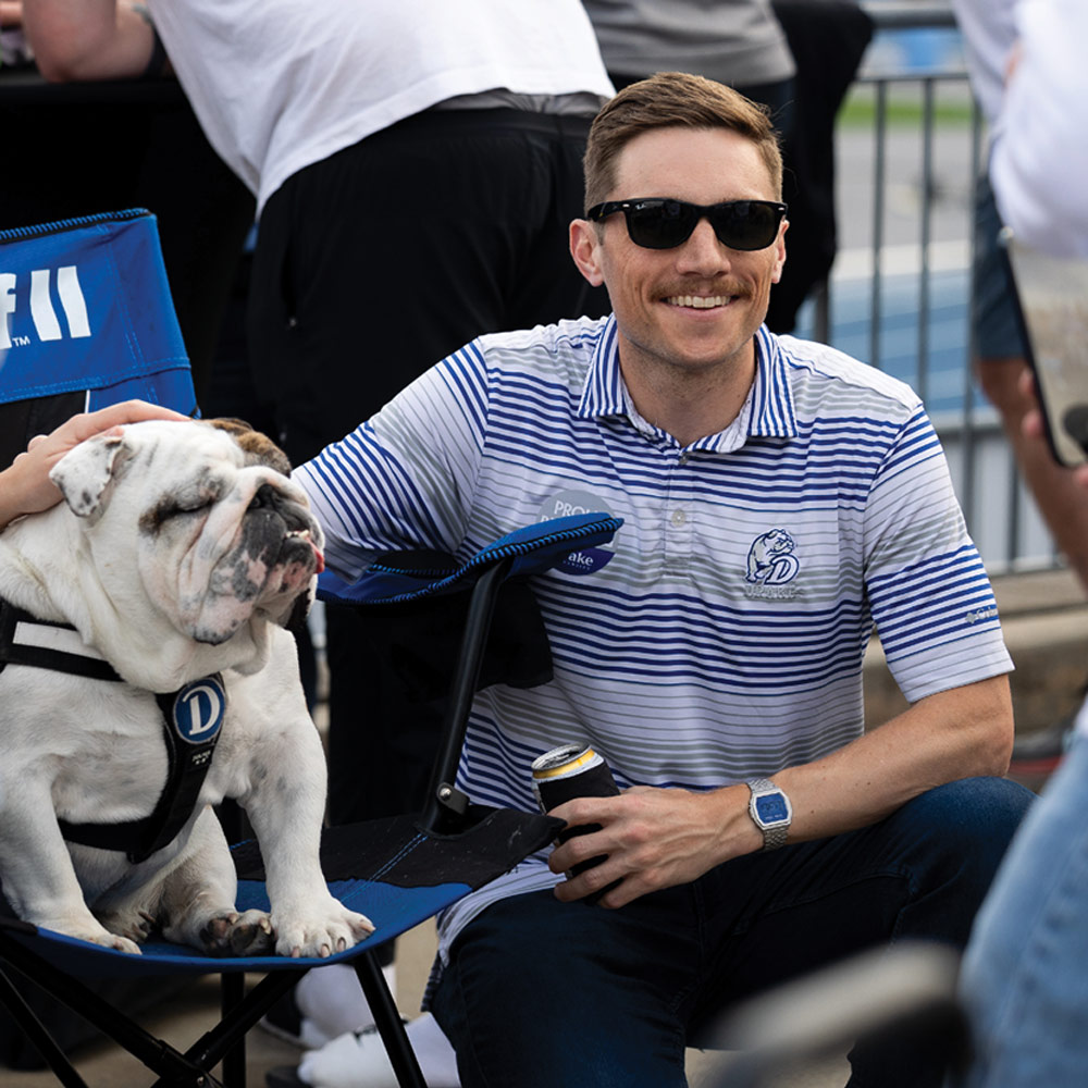 A man with a mustache wearing sunglasses and a striped Drake polo shirt sits in a folding chair next to a bulldog mascot. He is holding a beverage can.