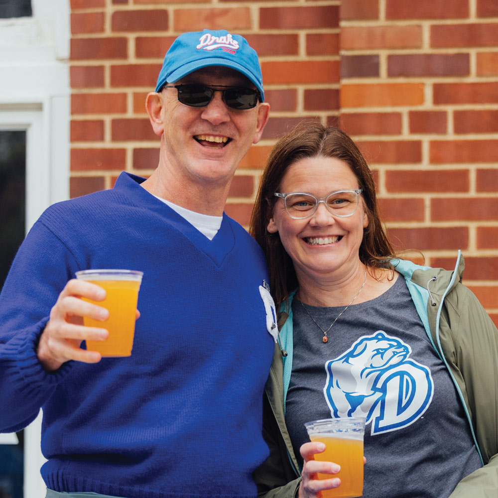 A man in a blue sweater and a woman in a grey Drake bulldog t-shirt smile while holding clear plastic cups of beer at an outdoor event.