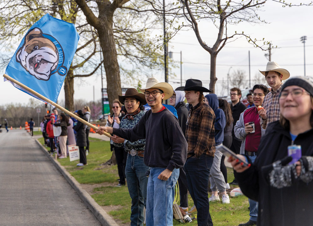 A group of spectators, including some in cowboy hats, cheer along a road. One man waves a light blue flag featuring a bulldog mascot during the Drake Relays event.