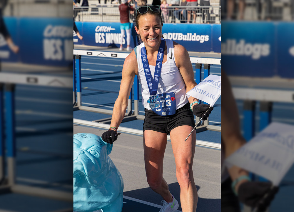 A smiling female runner wearing a 2025 Half Finisher medal and bib 73 poses on a track next to a blue bulldog statue, holding a "Champion" flag.