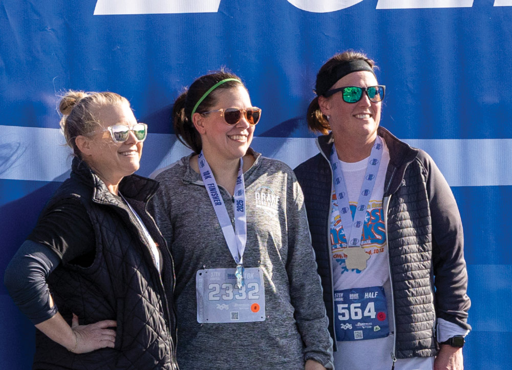 Three women wearing race medals and sunglasses pose together in front of a blue Drake Relays backdrop. The center runner wears bib 2332; the right wears bib 564.