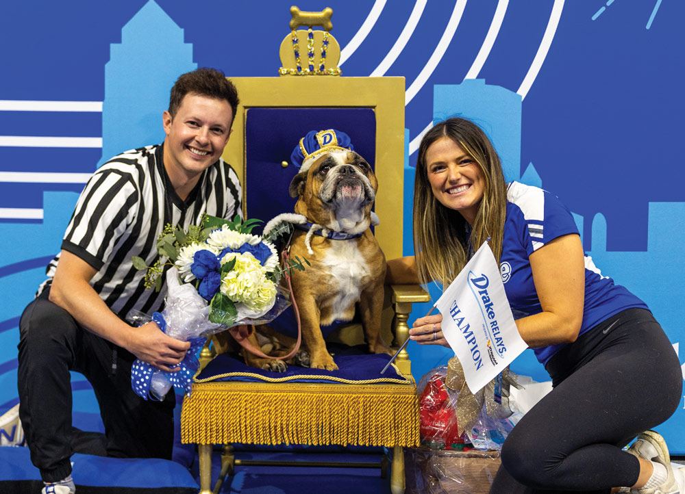 Two people kneel beside a bulldog wearing a blue and gold crown and cape, seated on a gold throne. A "Drake Relays Champion" flag and flowers are displayed.