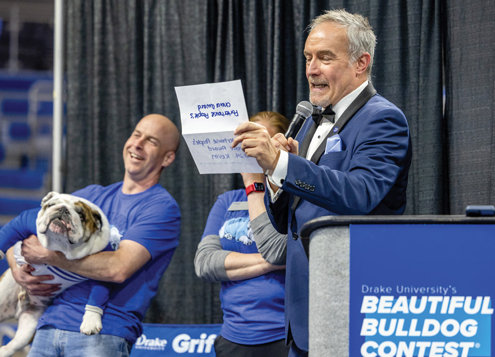 A man in a blue tuxedo speaks into a microphone at a podium for the "Beautiful Bulldog Contest" while holding a paper. A man in the background holds a bulldog.