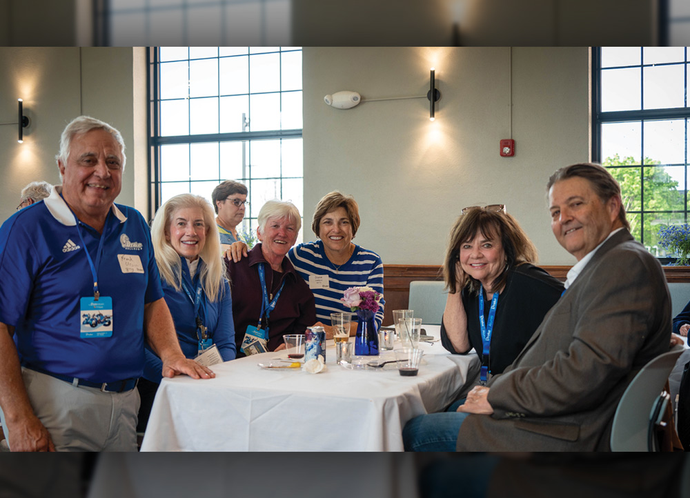 A group of six alumni pose for a photo while seated around a white cloth dinner table at an indoor banquet. They are wearing blue Drake University lanyards.