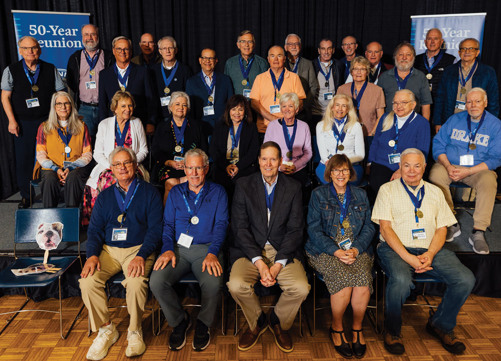 A group portrait of approximately 25 Drake University alumni posing at their 50-Year Reunion. Most are wearing blue lanyards and commemorative medals while sitting or standing on a stage.