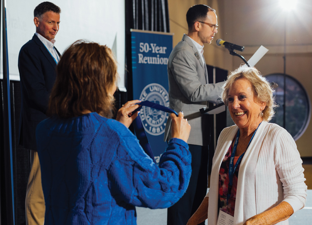 A woman in a white cardigan smiles as someone prepares to place a reunion medal around her neck. In the background, a man speaks at a podium marked with the Drake 50-Year Reunion logo.