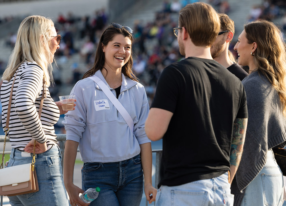 A young woman named Isabelle Tharp laughs while talking to a group of friends outdoors. She is wearing a light blue quarter-zip pullover and has sunglasses resting on her head.