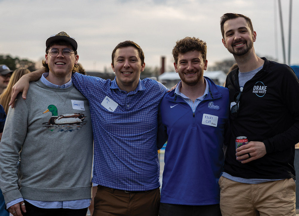 Four young men with arms around each other's shoulders pose for a photo at an outdoor event. One wears a grey sweater with a duck graphic, and another wears a black "Drake Road Races" shirt.