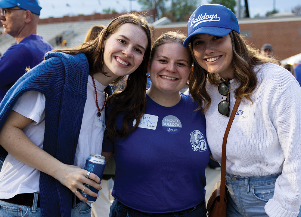 Three smiling young women pose together outdoors. They are wearing casual Drake apparel, including a blue "Bulldogs" hat and "Proud Bulldog" stickers. One holds a blue beverage can.