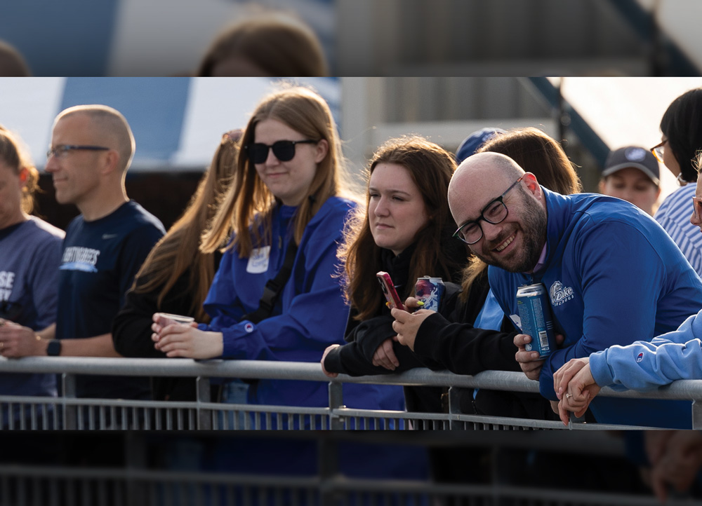 Spectators lean against a metal railing at an outdoor venue. A man in a blue Drake quarter-zip smiles at the camera while holding a blue beverage can alongside other attendees.