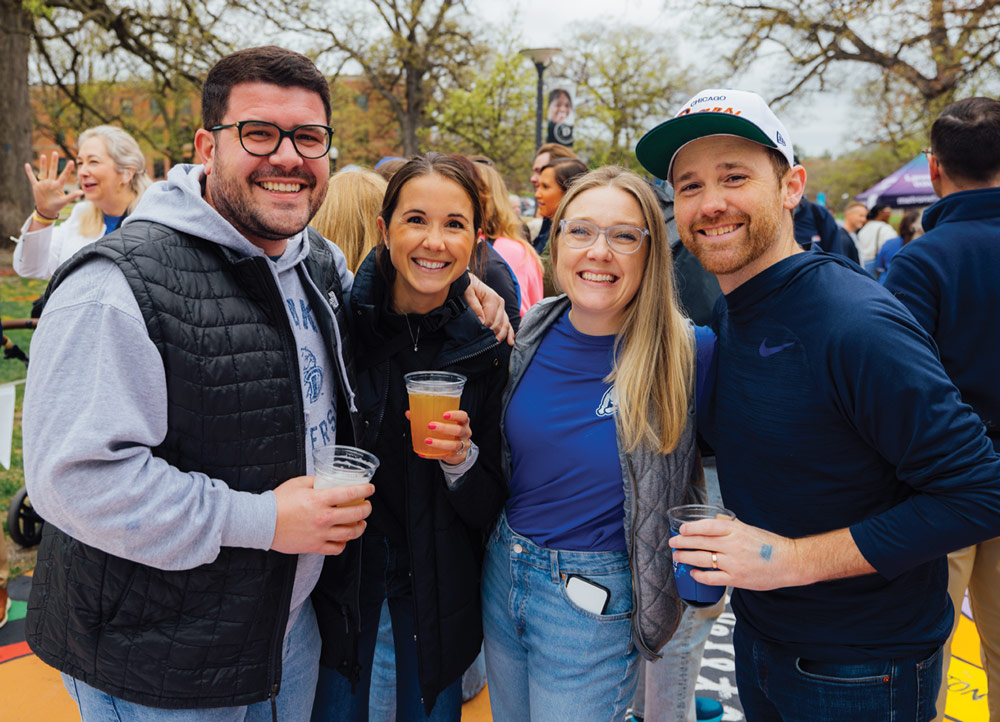 Two men and two women pose with plastic cups of beer at an outdoor alumni event. They are dressed in casual layers, including vests and hoodies, with a Drake University logo visible.