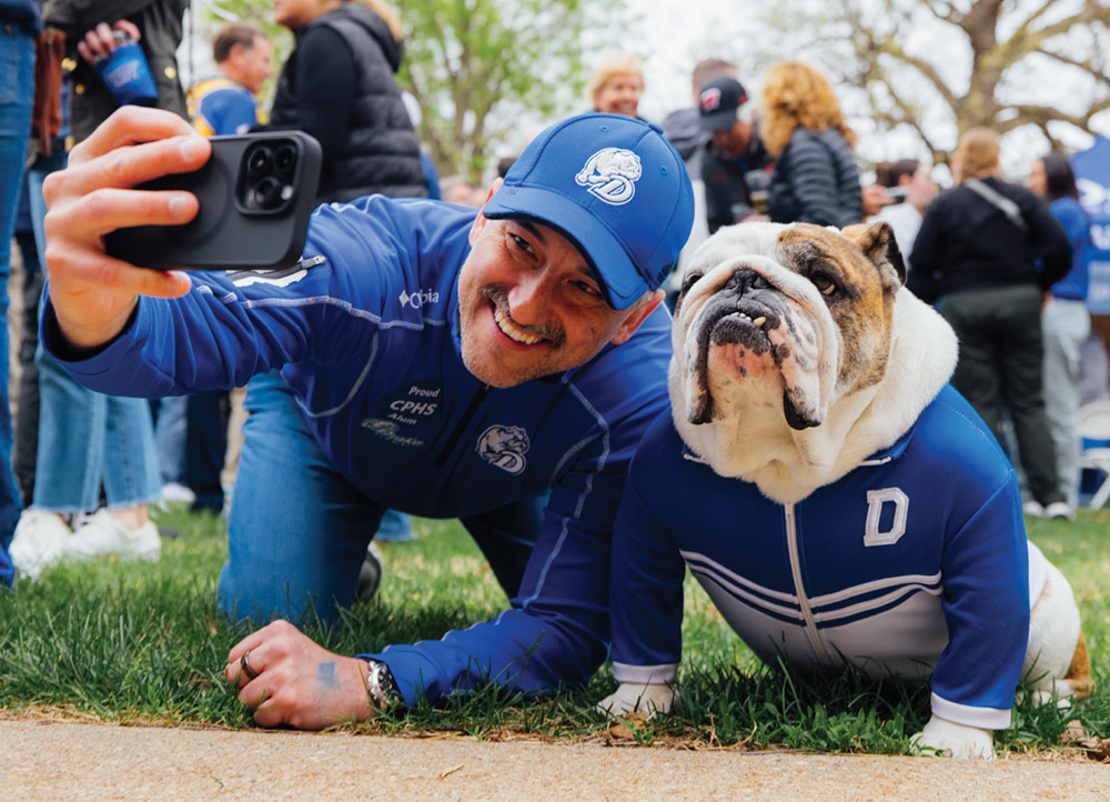 A man wearing a blue Drake University cap and pullover kneels on the grass to take a selfie with a bulldog. The bulldog is wearing a matching blue and white Drake athletic jacket.