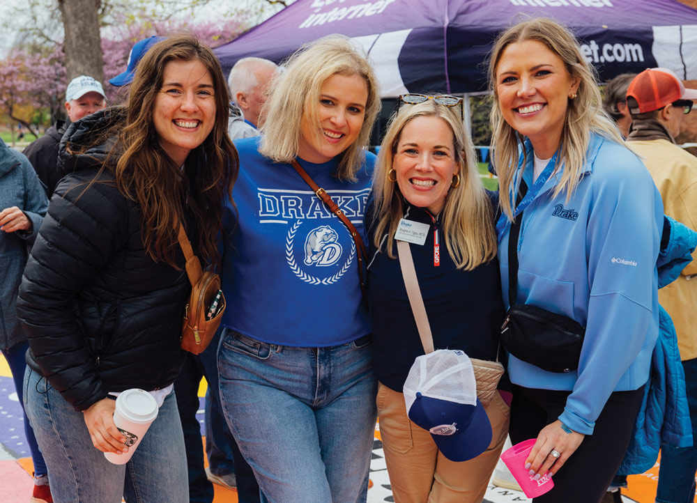 Four women smile together at an outdoor event near a purple tent. They are wearing Drake apparel, including a blue sweatshirt with the bulldog mascot and a light blue Columbia jacket.