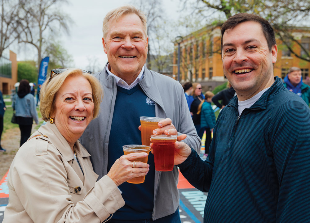 Two men and one woman smile for a group photo outdoors while holding plastic cups of beer. They are dressed in casual layers, including a tan trench coat and a grey zip-up jacket.