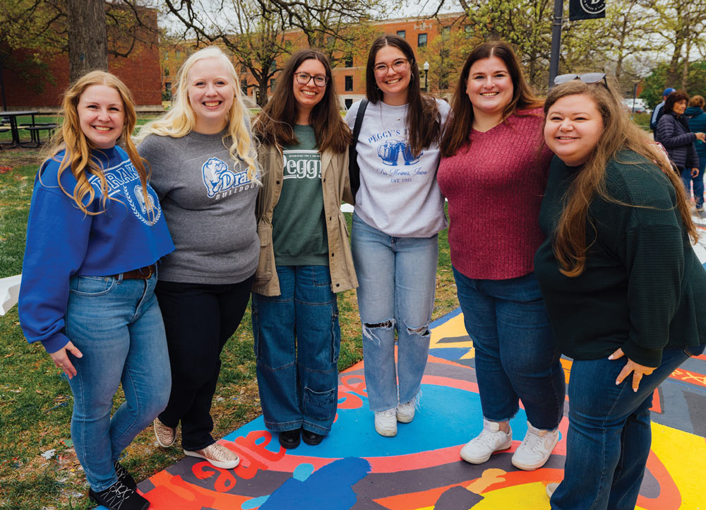 Six young women stand in a line on a colorful painted pavement, smiling for the camera. They wear casual Drake University apparel and jeans while posing outdoors on a cloudy day.