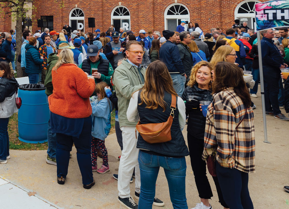 A large crowd of people gathers outdoors in front of a brick building for a social event. Attendees are seen talking, drinking from plastic cups, and wearing casual spring jackets.