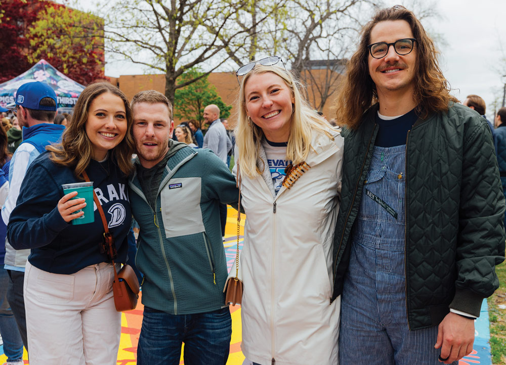Four friends, two men and two women, pose together at an outdoor event. One woman holds a green plastic cup, and one man wears blue striped overalls under a quilted olive green jacket.