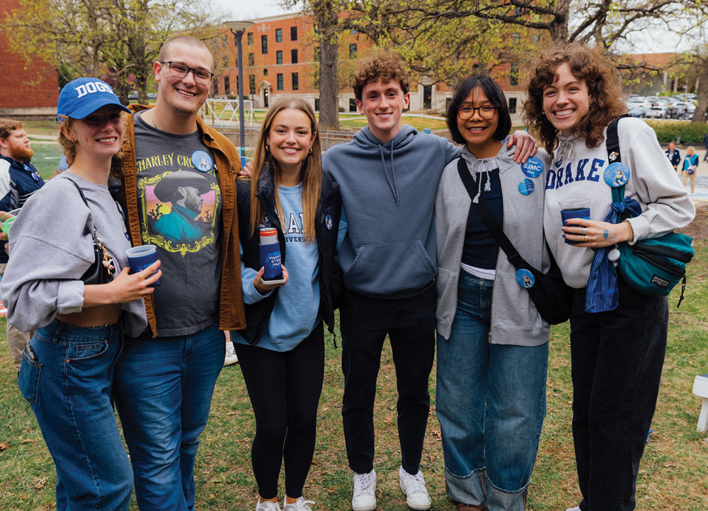 Six young adults stand together on a lawn, smiling for a group portrait. They wear casual clothing, including a blue "Dogs" hat, a grey hoodie, and a sweatshirt with a cowboy graphic.
