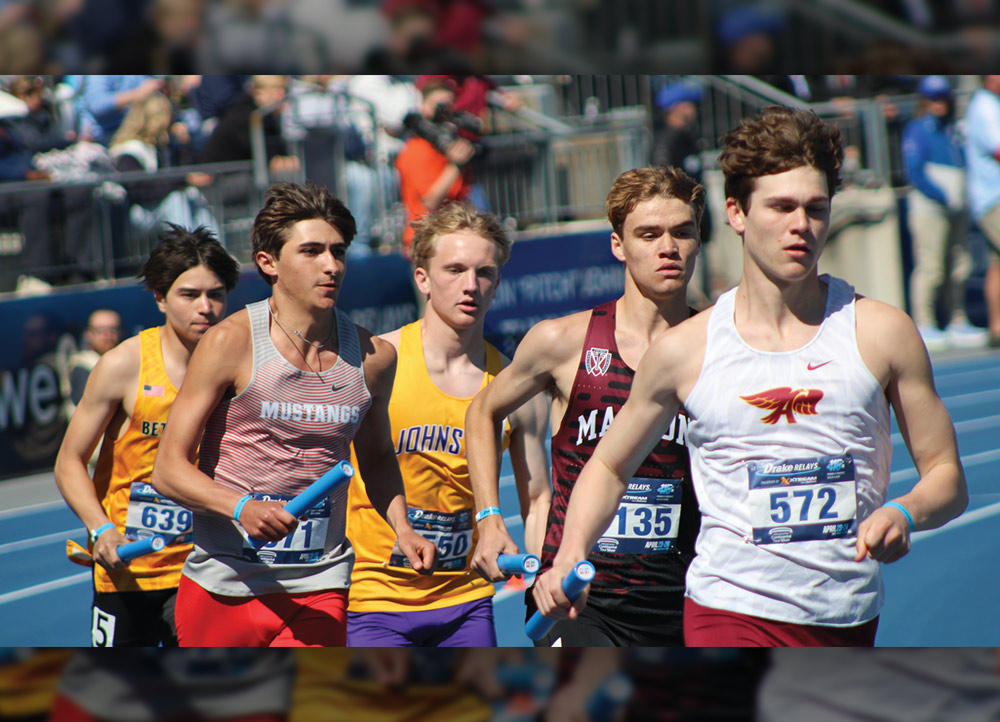 Five male track athletes compete in a relay race on a blue track. The runners, wearing various school singlets and holding blue batons, are focused as they sprint toward the camera.