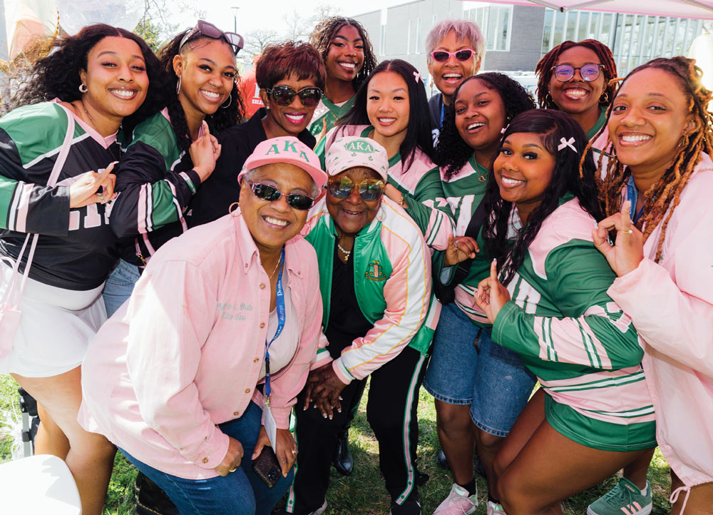 A large group of women wearing green and pink Kappa Alpha Theta sorority apparel smile for a close-up photo outdoors. Many are holding up a sorority hand sign and wearing sunglasses.