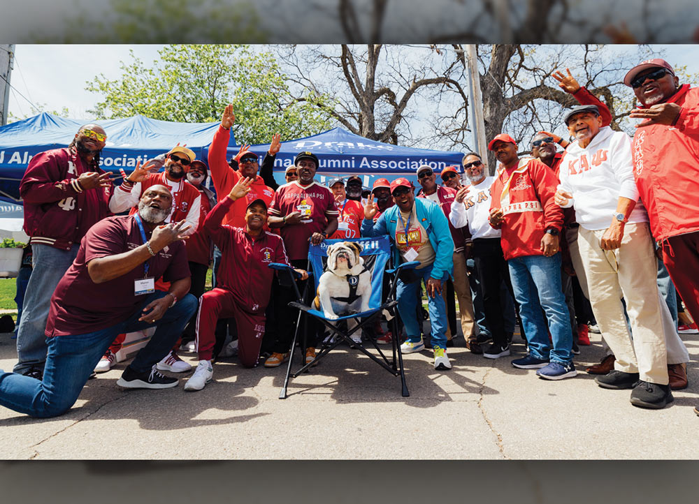 A group of men wearing Kappa Alpha Psi fraternity apparel gather around a bulldog mascot sitting in a blue chair. Many are wearing red jackets or hats and making fraternity hand signs.
