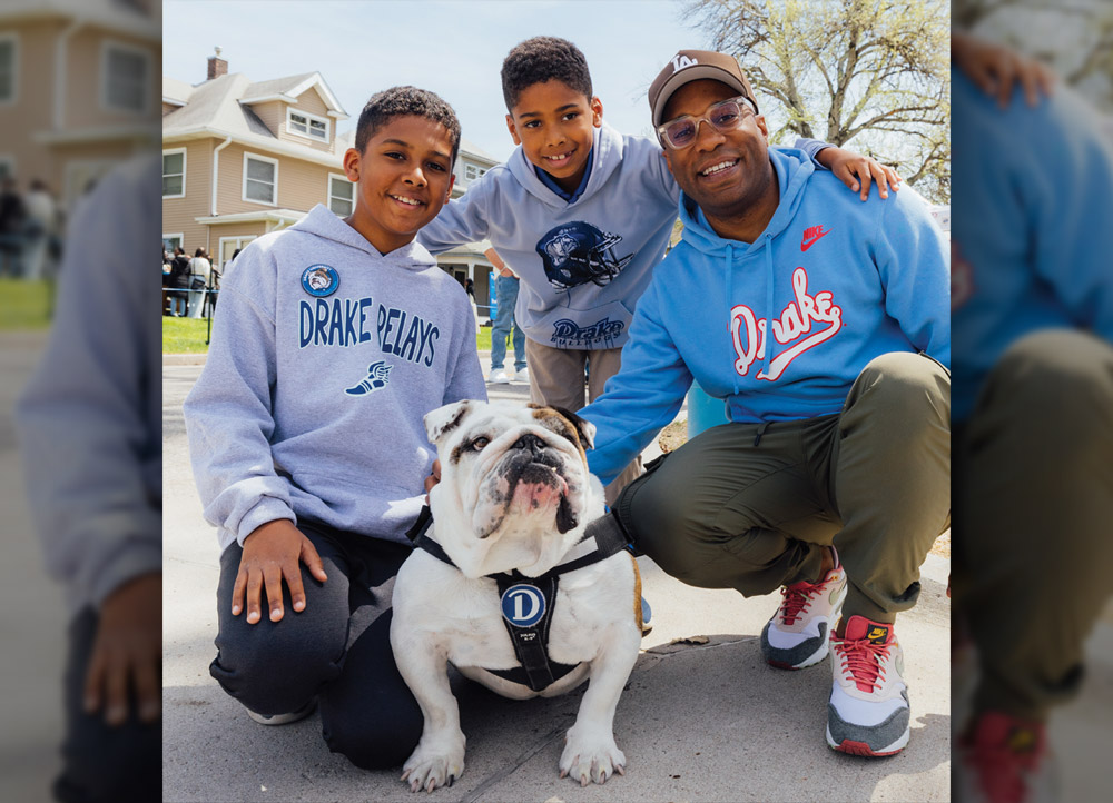 A man and two young boys kneel for a photo with a bulldog mascot on a sidewalk. They wear "Drake Relays" and "Drake Bulldogs" hoodies in various shades of blue and grey.
