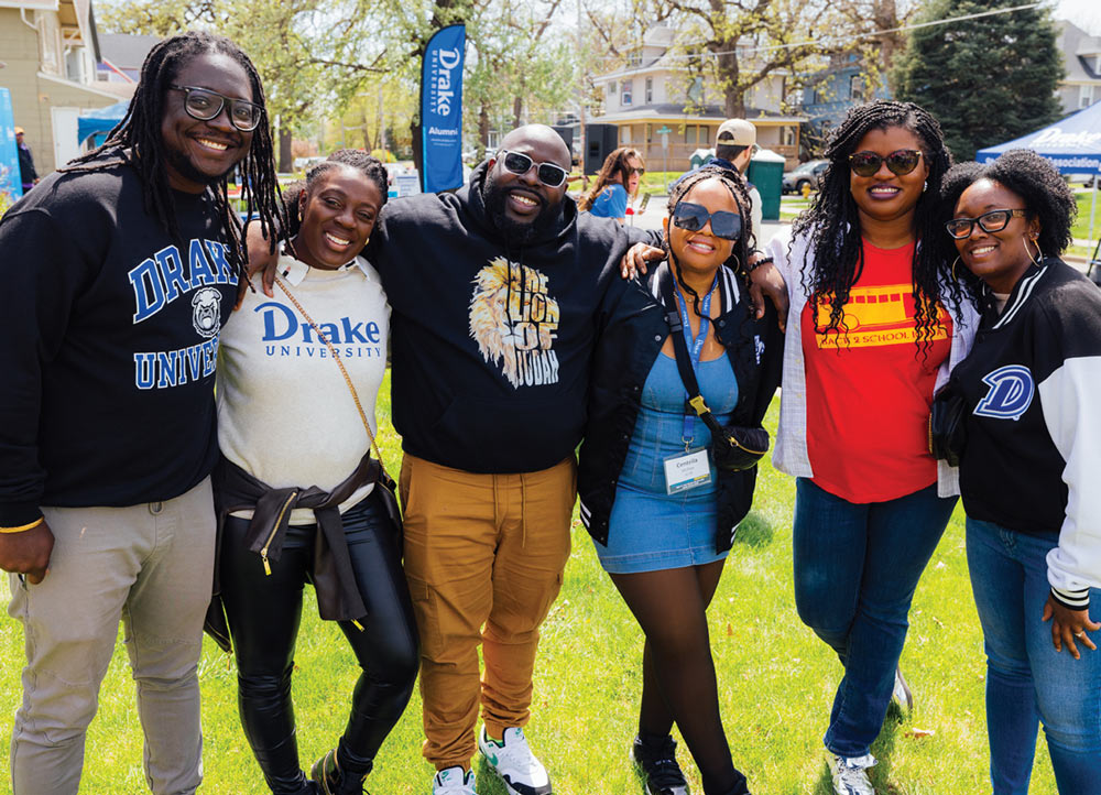 Six alumni pose together on a lawn, smiling for the camera. They wear various Drake University apparel, including a black "Drake University" sweatshirt with a bulldog logo.