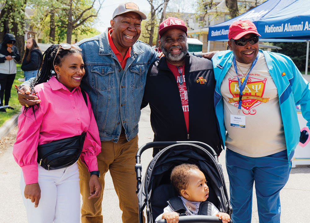 A group of four adults, three men and one woman, smile and pose together outdoors behind a baby in a black stroller. Two men wear red "Kappa Alpha Psi" apparel.