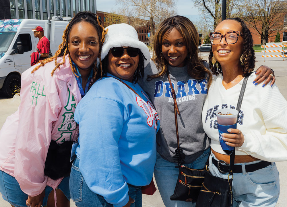 Four women smile together outdoors. One wears a pink Alpha Kappa Alpha jacket; another wears a grey "Drake Bulldogs" sweatshirt. They are dressed in casual jeans and layers.