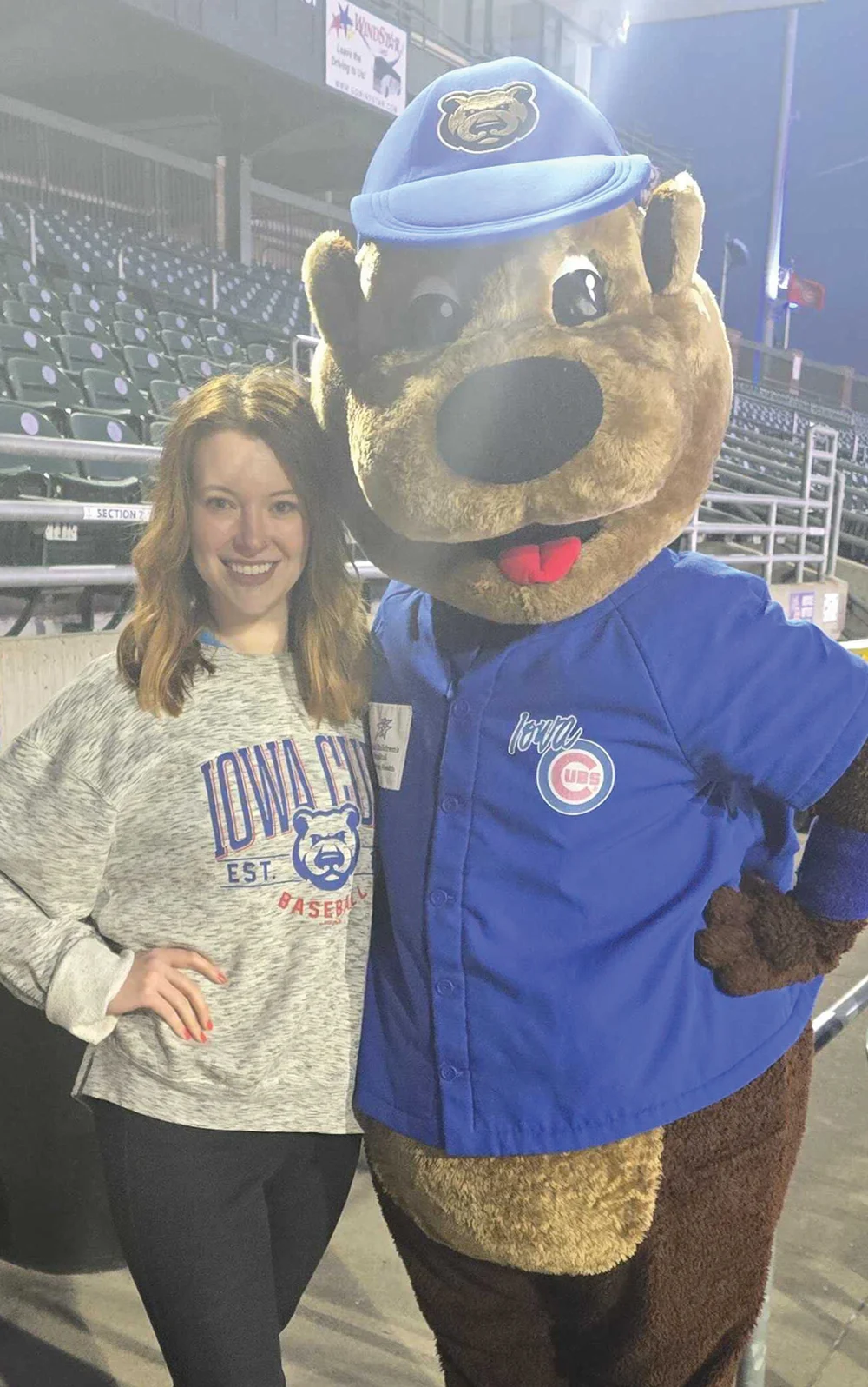 A smiling woman in a gray Iowa Cubs sweatshirt poses next to the Iowa Cubs bear mascot in a blue jersey at a stadium at night.