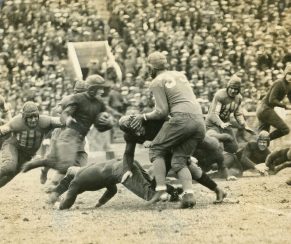 Grainy, vintage action shot of a football game with players in leather helmets and padded jerseys clashing on a grass field in front of a blurred crowd.