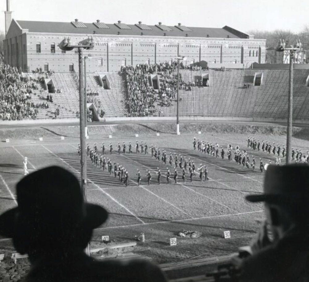 Vintage black and white photo from the stands showing a marching band in formation on a football field in front of a large brick fieldhouse and crowded bleachers.