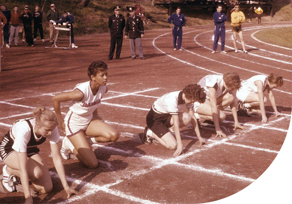 Historic color photo of five female track athletes in white uniforms crouched in starting blocks on a red dirt track, ready to begin a race.