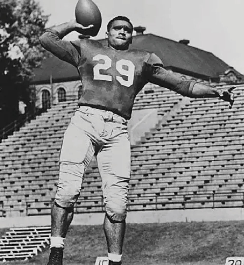 Black and white portrait of a football player in jersey number 29 preparing to throw a pass, with the stadium bleachers and fieldhouse in the background.