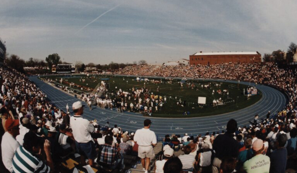 Wide-angle view of a crowded stadium with a blue track under a clear sky; officials and athletes gather on the central grass field near a large brick building.